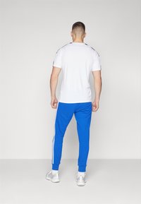 White short-sleeve T-shirt with black accents, paired with blue track pants featuring white side stripes. Sports shoes in light gray.
