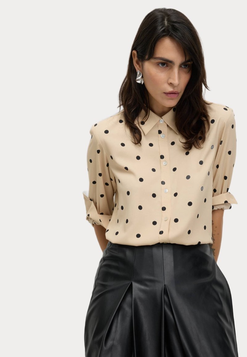 Beige blus med prickar, knäppning fram och uppvikta ärmar, i kombination med en svart plisserad läderkjol.