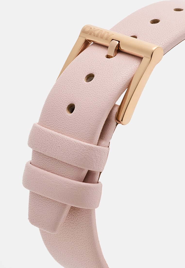 Jasnoróżowy pasek zegarka z syntetycznej skóry z teksturowaną powierzchnią, z metalową klamrą w kolorze złotym i kilkoma otworami do regulacji.