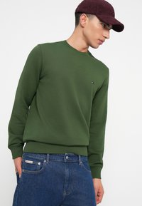 Egy férfi zöld környakú pulóverben, kék farmernadrágban és bordó sapkában jobbra néz, az egyik keze a zsebében van.