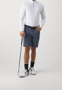 Hombre vestido con camisa de manga larga blanca y pantalones cortos marinos con patrón de logo de Calvin Klein, sosteniendo un palo de golf, de pie sobre un fondo liso.