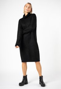 Strickkleid in Schwarz mit hohem Schalkragen, langen Ärmeln und lässiger Passform, ausgestattet mit geripptem Muster und knieumspielender Länge. Schwarze Knöchelstiefel.