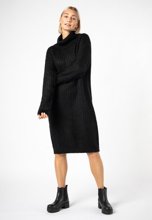Strickkleid in Schwarz mit hohem Schalkragen, langen Ärmeln und lässiger Passform, ausgestattet mit geripptem Muster und knieumspielender Länge. Schwarze Knöchelstiefel.
