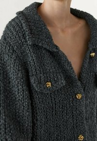 Cardigan lavorato a maglia grigio spesso con scollo a collo, caratterizzato da bottoni dorati e due tasche sul petto. Tessuto morbido e vestibilità ampia.