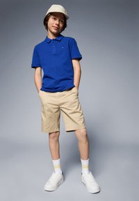 Blå polo skjorte, khaki shorts, hvite joggesko, sokker med gule striper og en lys caps. Avslappet antrekk på en ungdom.
