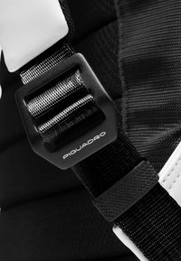 Zwarte verstelbare riem met een textuur, voorzien van een zwarte kunststof gesp met het opschrift "Piquadro" en een wit gewatteerd accent in de buurt.