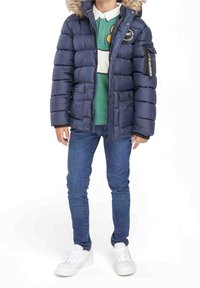 Dunjacka i marinblått med en huva i fuskpäls, dragkedja och tryckknappar, samt patchdetaljer. Bärs över en grön polo och blåjeans.