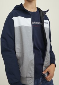 Färgbelagd jacka med sektioner i marinblått, grått och vitt. Har en huva, dragkedjestängning och framfickor. Mjuk tygtextur.