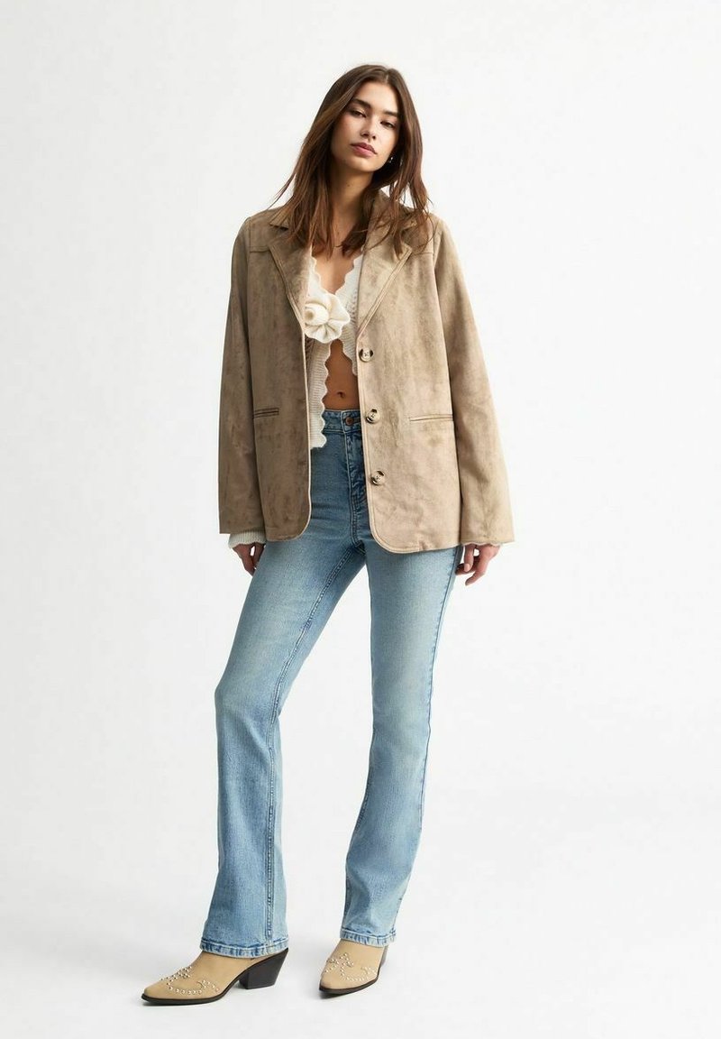Blazer de ante beige con un corte relajado, gran cuello y cierre de botón, combinado con unos vaqueros de campana azul claro y zapatos de tacón beige.