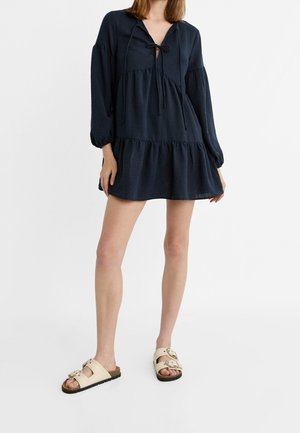 Femme portant une robe ample bleu marine à manches longues, à col noué, et des sandales beige à boucle, debout sur un fond blanc.