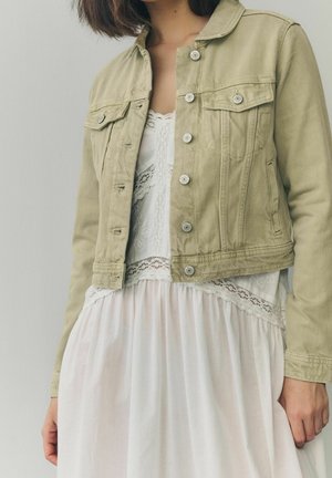 Woman wearing a beige denim jacket over a white lace-trimmed dress standing against a plain light background.
