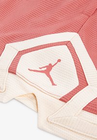 Síťované sportovní šortky v korálové a krémové barvě. Mají texturovanou tkaninu, výrazné logo v korálové barvě a kontrastní lemování podél okrajů.