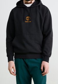 Sudadera con capucha negra hecha de una mezcla de algodón, con un bolsillo frontal y un logo de Timberland bordado en marrón. Con puños y dobladillo acanalados.