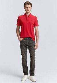 Polo rouge en tissu doux, associé à un jean gris foncé. Le mannequin porte des baskets blanches. Design simple avec un col classique.