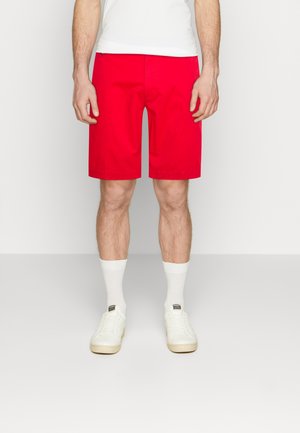 Man wearing bright red knee-length shorts, white socks, white sneakers, and a white shirt, standing against a plain background.
