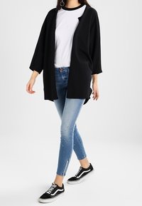 Svart öppen kavaj över en vit t-shirt, ihopparad med blå skinny jeans med en lätt slitdetalj och svarta sneakers.