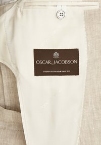 Beige tyg med en texturerad brun etikett som läser "OSCAR JACOBSON" och "SVENSK HANTVERKSKUNSKAP SEDAN 1903," med en knapp och sömdetaljer.