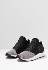 Mustat ja valkoiset liukuesteet, joissa on tekstuurilla varustettu kärki, joustava nauhoitus ja valkoiset pohjat, valkoisella pinnalla, yksinkertaisen taustan kanssa.