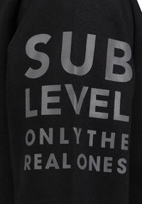 Zwarte hoodie met gedurfde, zilveren tekst "SUB LEVEL ONLY THE REAL ONES" prominent afgebeeld. Katoenen materiaal met een gladde textuur.