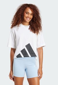 Bela skrajšana majica s črnim grafičnim logotipom Adidas, v kombinaciji s svetlo modrimi športnimi kratkimi hlačami z belimi stranskimi črtami in majhnim logotipom.