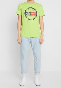 Zielona bawełniana koszulka z okrągłym nadrukiem graficznym z napisem "TOMMY JEANS", w zestawieniu z jasnoniebieskimi dżinsami i białymi sneakersami.