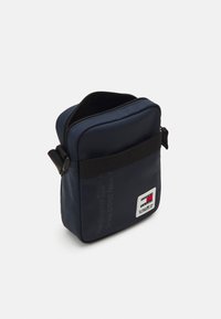 Borsa a tracolla in nylon blu navy con chiusura a zip, tracolla regolabile e patch con logo sul fronte. Tessuto liscio e forma rettangolare.