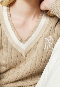 Beige kabelstickad tröja med vit V-ringning och broderad logotypdetalj på vänster bröst. Texturen är mjuk och ribbad.