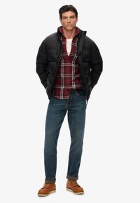 Chaqueta de abrigo negra, camisa a cuadros roja y negra, jeans azules y botas marrones. La chaqueta tiene un cuello alto; la camisa presenta detalles de botones.