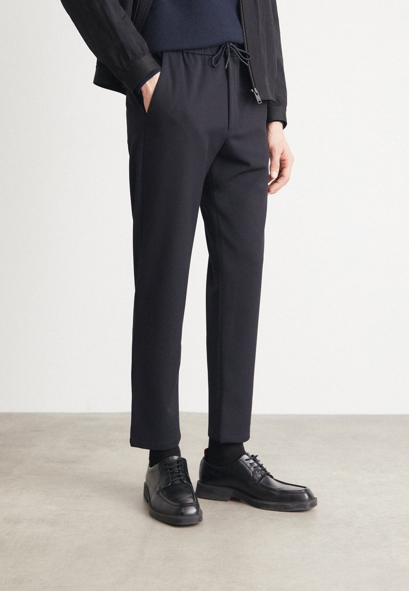 Personne portant un pantalon noir à cordon, des chaussettes noires et des chaussures en cuir noires, debout sur un sol gris clair.