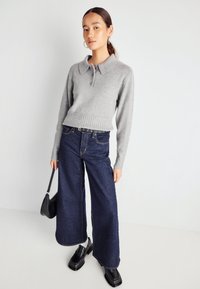 Siv brezast pulover z ovratnikom, usklajen s širokimi temno jeans hlačami in črnimi debelimi čevlji. Model drži majhno črno torbico.