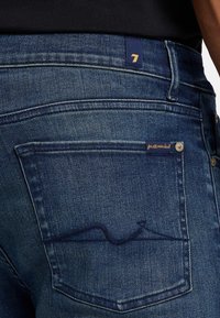 Ciemnoniebieskie dżinsy z charakterystycznym wzorem tylnych kieszeni, kontrastowym przeszyciem oraz małą metką z logo blisko pasa.