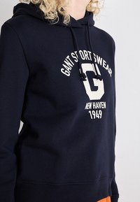 Personne portant un sweat à capuche bleu marine avec le texte blanc "GANT SPORTSWEAR", un grand "G" et "NEW HAVEN 1949" à l'avant.