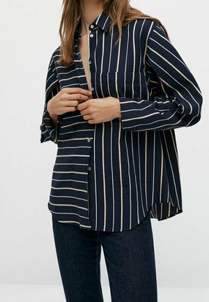 Person buttoning a loose-fitting navy blue shirt with white vertical stripes, paired with dark blue pants against a plain background.