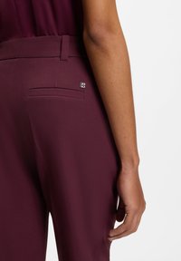 Burgundy trousers with a smooth fabric, featuring a back pocket and a small metallic accent near the waistband. Tailored fit.