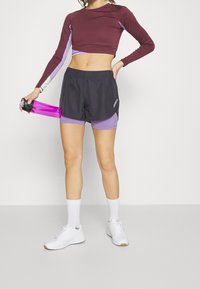 Maroninpitkähiha crop-top, jossa on violetteja yksityiskohtia, yhdistetty mustiin shortseihin ja violettiin leggingiin. Pitelee vaaleanpunaista vesipulloa; valkoiset kengät.
