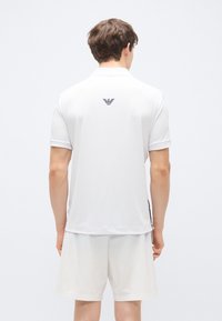 Weißes Polohemd mit kurzen Ärmeln und einem schwarzen Logo im Nackenbereich. Der Stoff wirkt glatt; getragen mit passenden weißen Shorts. Minimalistisches Design.