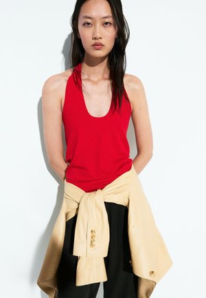 Femme aux longs cheveux foncés portant un top rouge sans manches et une veste beige nouée autour de sa taille sur un fond blanc uni.