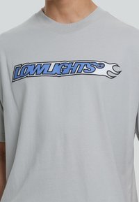 Jasnoszary bawełniany t-shirt z niebiesko-czarnym haftowanym logo "LOWLIGHTS" o stylizowanych brzegach oraz okrągłym dekoltem.