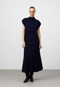Donna in un top senza maniche blu navy e una gonna lunga abbinata, che indossa stivali neri con punta su uno sfondo chiaro e semplice.