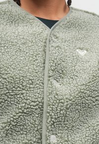 Zelený fleecový kardigán s textúrovaným, nadýchaným povrchom, sivým lemovaním a patentkami. Na ľavej strane je malý biely logotyp.