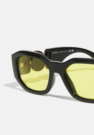 Óculos de sol oversized pretos com armação geométrica, apresentando lentes amarelas e detalhes dourados nas dobradiças. Textura mate estilosa.