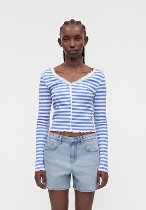 Kobieta ubrana w niebiesko-biały paskowany, długi rękaw, zapinany na guziki crop top i jasnoniebieskie szorty jeansowe, stojąca na jednolitym tle.