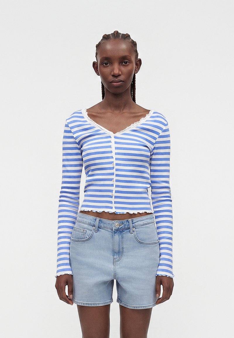 Γυναίκα που φοράει μπλε και λευκό ριγέ crop top με κουμπιά και μακριά μανίκια και ανοιχτά μπλε τζιν σορτς, που στέκεται σε απλό φόντο.