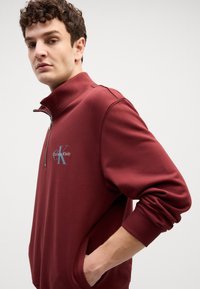 Βουργουνδί κραγιόν-zip φούτερ με ριγέ μανσέτες, διαθέτοντας ένα γκρι λογότυπο Calvin Klein στο στήθος και χαλαρή εφαρμογή. Μαλακή υφή υφάσματος.