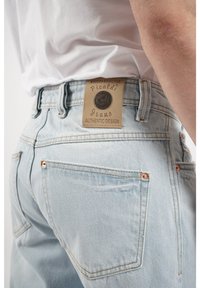 Jeans de mezclilla azul claro que presentan una etiqueta beige con un logo en relieve, remaches de cobre y costuras a tono en los bolsillos traseros.