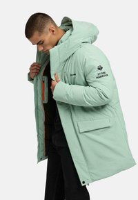 Jasnozielona kurtka puchowa z dużym kapturem, zamek błyskawiczny i boczne kieszenie; posiada czarne logo i pomarańczową podszewkę. Miękka, teksturowana tkanina.