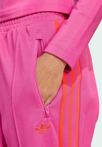 Roze sportieve trainingspak met ritszak, voorzien van oranje en paarse zijstrepen en een geborduurd logo. Materiaal lijkt glad en lichtgewicht.