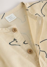 Beež beebiriietus pehme soonilise tekstuuriga, sinise loomakontuurse mustriga ja kahe puidust nööbiga kaelal. Silt nähtav.