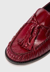 Mocassins de couro bordô com detalhes em franjas, apresentando uma ponta arredondada e superfície texturizada, contornados por uma sola de borracha preta.