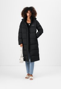 Black puffer coat with a hood, featuring horizontal quilting, front buttons, and a logo on the chest. Paired with light jeans and cream sandals.